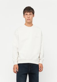 Sudadera de color beige claro con cuello redondo, puños y dobladillo de canalé. Presenta un sutil detalle de logo en el frente. Se lleva con jeans oscuros.