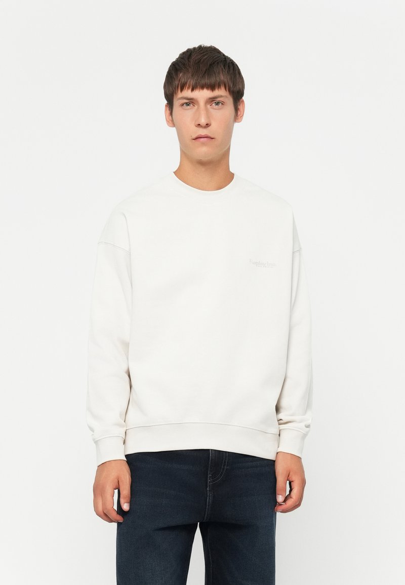 Sudadera de color beige claro con cuello redondo, puños y dobladillo de canalé. Presenta un sutil detalle de logo en el frente. Se lleva con jeans oscuros.