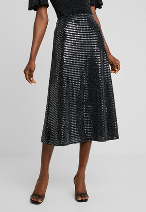 Sort palietbesat midi nederdel båret med sorte hælede sandaler på en person, der står mod en ensfarvet grå baggrund.