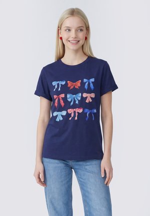 Blonde vrouw met rode oorbellen en een marineblauw T-shirt met negen rode, witte en blauwe strikjes, gecombineerd met lichtblauwe spijkerbroek.