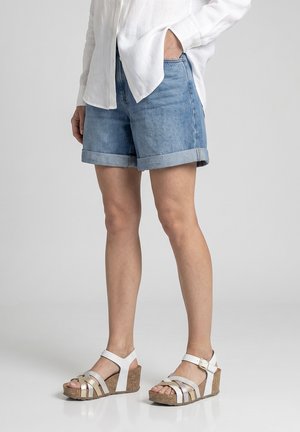 Persona que lleva shorts vaqueros azules con puños, camisa blanca de manga larga con botones y sandalias de cuña blancas con suelas de corcho.
