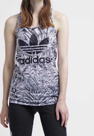 Kobieta ubrana w bezrękawnik Adidas z czarno-białymi liściastymi wzorami i czarne legginsy na jednolitym tle.