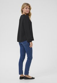 Haut noir à manches longues avec des épaules bouffantes, associé à un jean skinny bleu et des ballerines noires, vu de côté sur un fond neutre.