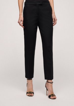 Fekete, slim fit szabású nadrág, lapos elülső résszel, oldalzsebekkel és sima textúrával. Fekete magassarkú szandállal viselve.