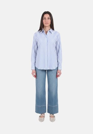 Donna in piedi di fronte, indossa una camicia a righe verticali blu e bianche con bottoni, jeans blu a gamba larga e ballerine bianche.