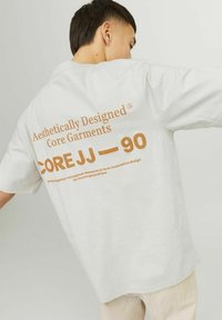 White short-sleeve T-shirt featuring large brown text on the back: "Aesthetically Designed Core Garments COREJJ—90," with curved neckline.
