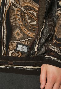 Sweter dzianinowy w ziemistych odcieniach z misternymi geometrycznymi wzorami, ściągaczami na mankietach oraz małą metką marki w srebrnym wykończeniu.