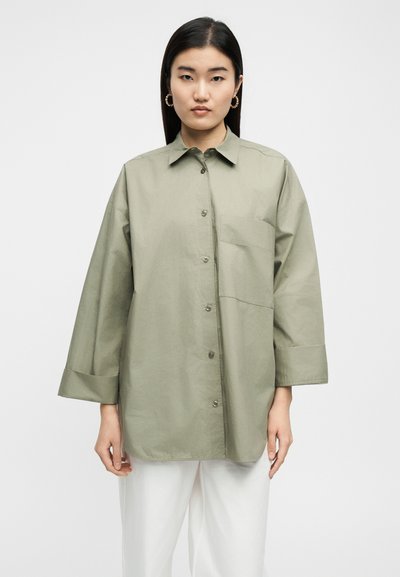 Γυναίκα με μακριά μαύρα μαλλιά που φοράει χρυσούς κρίκους στα αυτιά, φαρδιά γκρι-πράσινα πουκάμισα με κουμπιά και λευκό παντελόνι, στέκεται μπροστά σε απλό φόντο.