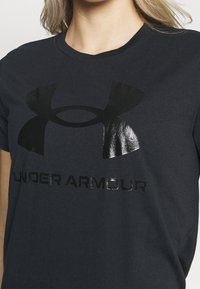 Kvinna som bär en svart Under Armour t-shirt med en stor blänkande logotyp och text på bröstet, beskuren under nacken.