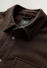 Chaqueta de camisa de corduroy marrón con un cuello pico, bolsillo frontal y cierre de botón oscuro. La etiqueta dice "NEXT". Detalle del tejido texturizado visible.