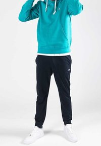 Turkos hoodie med kenguruficka och vita dragkedjor, kombinerat med mörka byxor och vita sneakers. Har små logotypdetaljer.