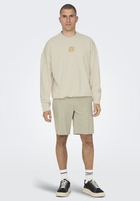 Beige sweatshirt med et guld logo, parret med lysegrønne shorts. Overtøjet er suppleret med sorte sneakers og hvide stribede sokker.