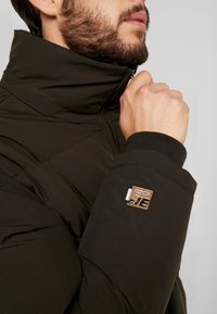 Mørkegrøn pufferjakke med høj krave, der har lynlåslukning, struktureret stof og et mærke med logo på venstre ærme.