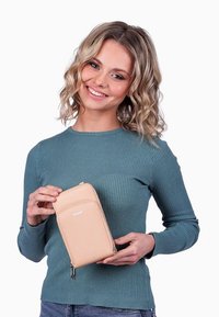 Béžová kožená peněženka se zipovým uzávěrem, štíhlý design a vytlačené logo. Model drží peněženku proti neutrálnímu pozadí.