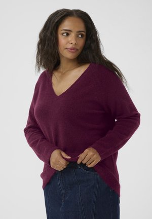 Burgundy v-neck sweater with long sleeves, made of soft material, featuring a relaxed fit and side slits, paired with a denim skirt.