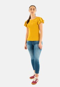Top de punto amarillo con canalé y mangas con volantes, combinado con jeans ajustados de color azul claro y zapatillas rojas con detalles blancos. Diseño simple y ajuste casual.