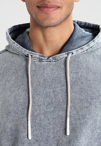 Nahaufnahme einer Person, die einen hellblauen, strukturierten Kapuzenpullover mit weißen Kordeln trägt, sichtbar sind der untere Teil des Gesichts und der Hals.