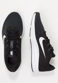 Des chaussures de sport noires avec une tige en mesh, un logo swoosh blanc, une semelle en caoutchouc et un système de laçage. Présente une surface texturée et un design de bout arrondi.