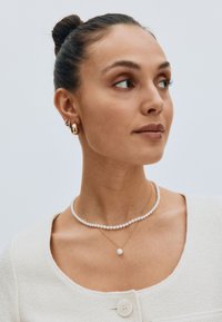 Pearl necklace and gold pendant on a woman with a textured beige top. Gold hoop and stud earrings. Hair in a bun. Neutral background.