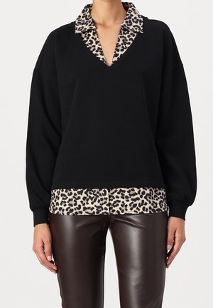 Femme portant un pull noir à col en V superposé sur une chemise à imprimé léopard avec col, associé à un pantalon en cuir marron foncé, debout devant un fond neutre.
