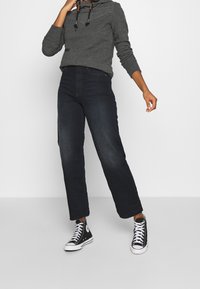 Persoon draagt een donkerblauwe rechte jeans, een grijze hoodie met lange mouwen en trekkoorden, en zwarte hoge sneakers met witte veters.