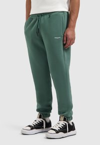 Groene katoenen joggers met een elastische tailleband, manchetten en een klein wit logo. Draag ze met zwart-witte sneakers. Soepele textuur.