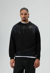 Zwarte sweatshirt met een gestructureerde "REBELLION" print in een druppelend ontwerp. Voorzien van een ronde hals en geribbelde manchetten, gecombineerd met donkere jeans.