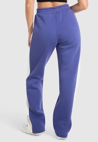 Pantalones deportivos de forro polar en color morado con paneles laterales blancos. Cintura elástica, diseño de pierna ancha y textura suave visible desde la vista trasera.