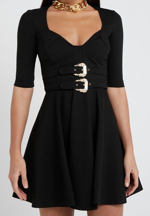 Frau trägt ein schwarzes Kleid mit ellbogenlangen Ärmeln, einem doppelt goldgeschnallten Gürtel und eine auffällige goldene Kette mit Löwenkopf-Anhänger.