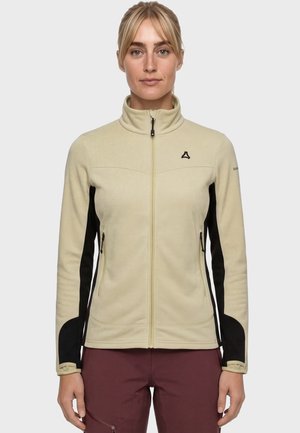 Frau trägt beige-schwarze Fleecejacke mit Reißverschluss und bordeauxrote Hose, steht vor schlichtem hellgrauem Hintergrund.