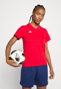 Červené športové tričko s výstrihom do V a krátkymi rukávmi, so vzorom bieleho loga Adidas, spojené s tmavomodrými športovými kraťasmi. Osoba drží futbalovú loptu s čierno-bielym vzorom.