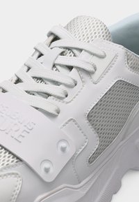 Κοντινή λήψη λευκού αθλητικού παπουτσιού με πάνελ από δίχτυ, χοντρές λευκές κορδόνια και μια λωρίδα με λογότυπο στο πάνω μέρος του παπουτσιού.