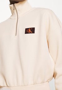 Beżowy rozpinany sweatshirt z wysokim kołnierzem, z prążkowanymi mankietami i dolną krawędzią. Posiada prostokątną czarną łatę z pomarańczowym logo "cK" na klatce piersiowej.