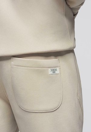 Pantaloni della tuta beige con una tasca posteriore che presenta un'etichetta bianca con la scritta "L'UNICO LIMITE SEI TU."