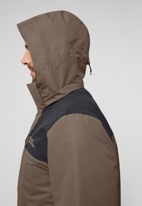 Bruine en zwarte waterdichte jas met een capuchon, voorzien van een ritssluiting en een strakke, gladde textuur. Het zijaanzicht toont een duidelijk kleurcontrast.