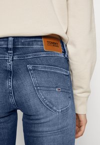 Jeans de mezclilla azul oscuro con bolsillos traseros, que cuentan con una etiqueta de cuero marrón de TOMMY JEANS y un pequeño logo de bandera en el bolsillo trasero.