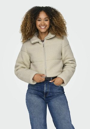Korte beige fleece jas met een rits aan de voorkant en hoge kraag. Heeft een gestructureerd oppervlak en zachte mouwen, gedragen over hoge taille jeans.