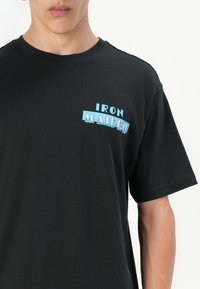 Sort bomulds t-shirt med "IRON MAIDEN" logo i blå og pink, placeret på venstre brystområde, rund hals og korte ærmer.