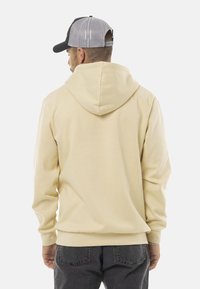 Haut à capuche beige avec un cordon de serrage, des poignets et un ourlet côtelés. Porté avec un jean gris foncé. La vue de dos montre une texture de tissu lisse.