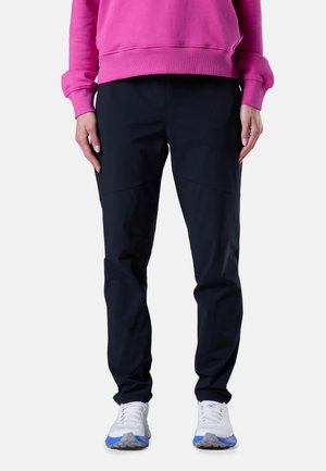 Osoba ubrana w czarne zwężane spodnie, różową bluzę z długim rękawem oraz białe trampki z niebieskimi akcentami, stojąca na jednolitym tle.