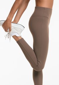 Hoch taillierte, braune Leggings aus elastischem Material mit nahtlosem Design und verstärkten Stitching, kombiniert mit weißen Sportschuhen.