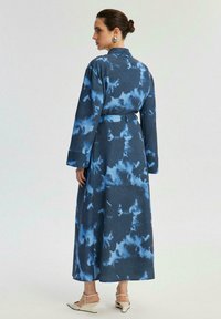 Pitkä, tummansininen takkimainen mekko, jossa on abstrakteja vaaleampia sinisiä kuvioita, vyötäröllä vyö ja leveät hihat. Tasainen pinta, nilkkapituisen muotoilu.