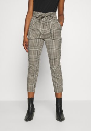 Femme portant un pantalon taille haute à carreaux noirs et blancs avec une ceinture nouée et des bottines noires, debout les mains dans les poches.