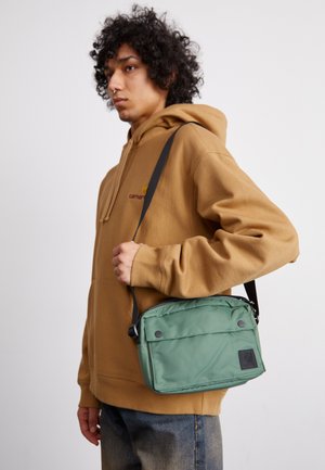 Zelená nylonová taška cez rameno so zipsovým uzáverom, nastaviteľným popruhom a predným klopovým vekom s cvokmi. Nosená osobou v hnedej mikine.