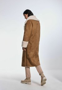 Brązowy płaszcz z faux zamszu z białym kołnierzem i mankietami z owczej skóry, o prostym kroju i średniej długości, zestawiony z beżowymi spodniami i botkami.