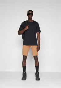 Svart kortärmad t-shirt, beige cargoshorts, svarta strumpor och vandringskängor. Modellen bär stora svarta solglasögon och håller en svart ryggsäck.