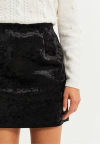 Svart sammetkjol med en slät yta och en åtsittande form, med sidofickor och en ren fåll, ihop med en texturerad vit tröja.