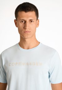 Ljusblå crew-neck t-shirt gjord av mjukt material, med den präglade texten "COPENHAGEN LINDBERGH" i beige färg över bröstet.
