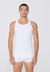 Débardeur en coton blanc avec de larges bretelles, associé à des culottes blanches. Le tissu semble lisse et extensible, épousant le corps du mannequin.