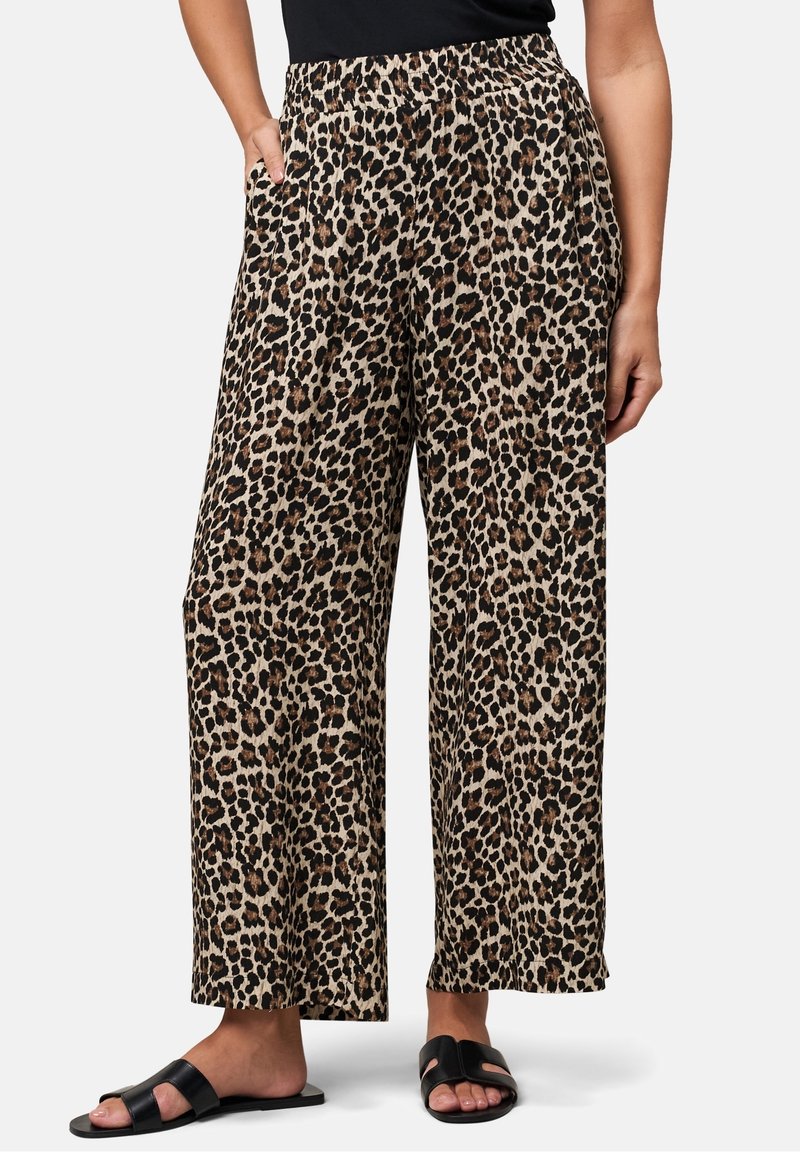 Pantalon large et confortable avec imprimé léopard, porté avec des sandales noires à enfiler et un top noir, les mains du mannequin détendues le long du corps.